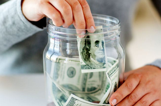 A person placing a $100 bill inside a jar half filled with cash