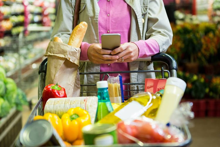 A woman using her phone while pushing a full shopping cart in a grocery store 
