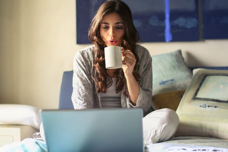 A woman sitting in bed with a laptop cooling off her coffee