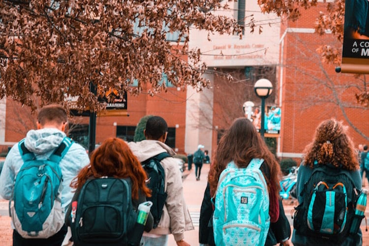 A group of students with blue backpacks walking toward a college building