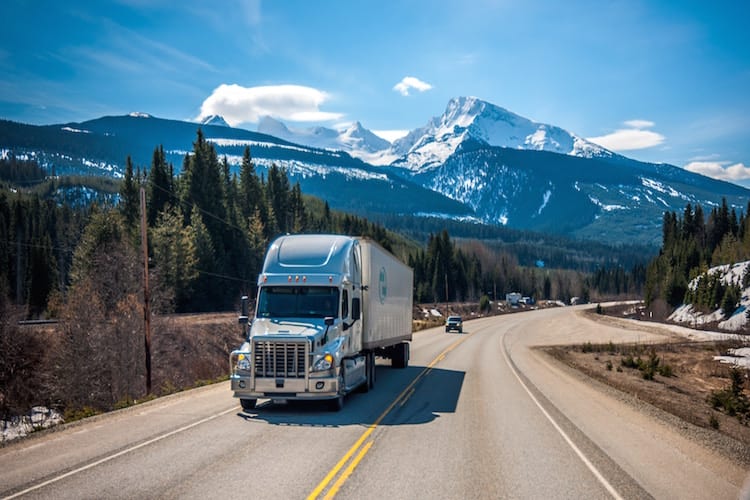 A cargo truck driving along a scenic highway with mountains and trees in the background