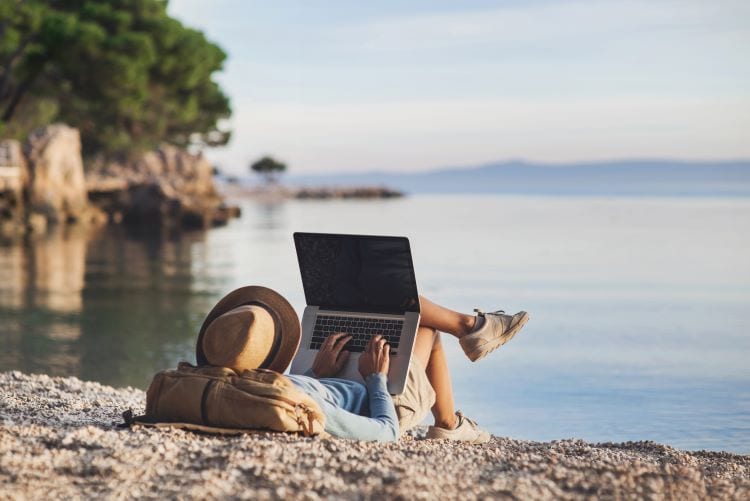 A person using their laptop on a beach with trees and water in front of them