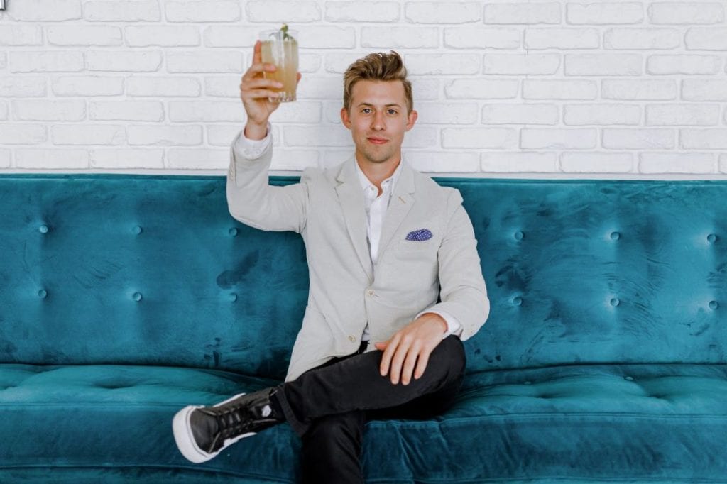 A man in a suit and sneakers on a blue couch raising a glass 