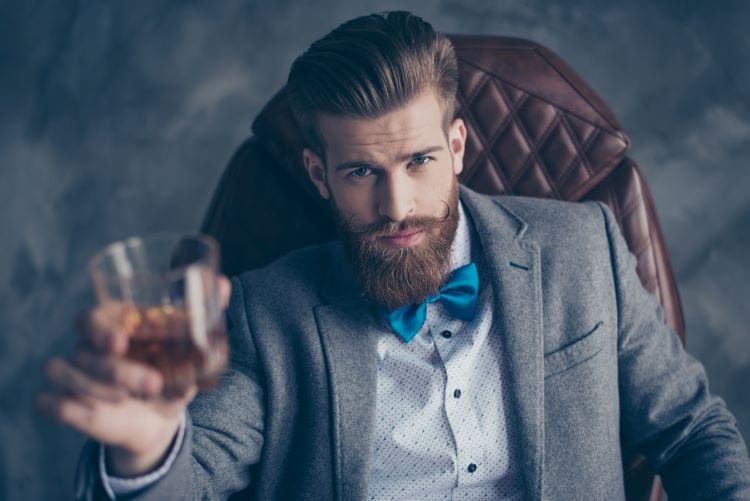A young man with a suit and beard holding a glass of liquor 