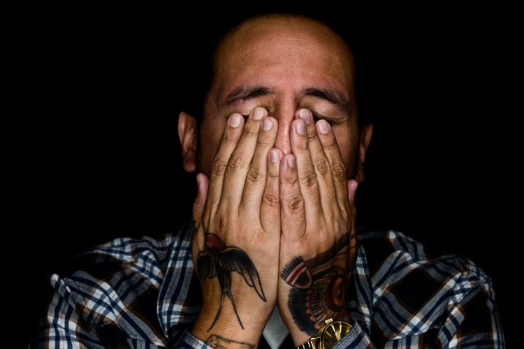 A man with tattoos on the backs of his hands covering his eyes and nose