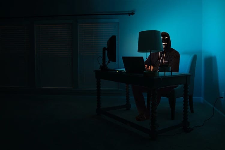 A person with a mask sitting in dim light behind a computer