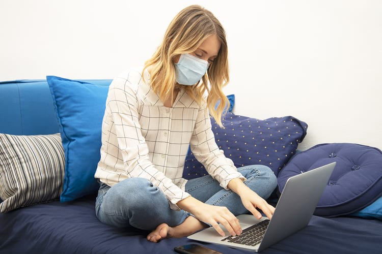 A woman on a couch wearing a mask using a laptop