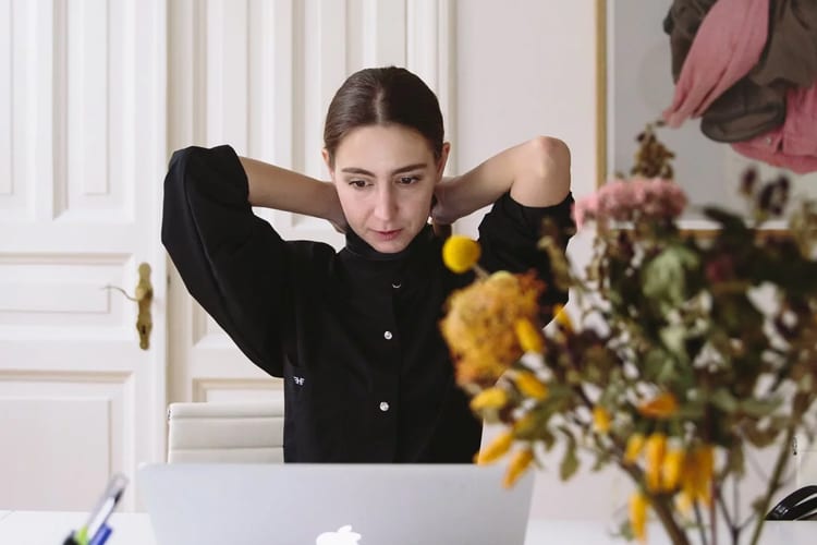 A brunette woman tying her hair back while working on her laptop