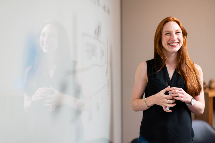 A woman with red hair standing next to a whiteboard and smiling