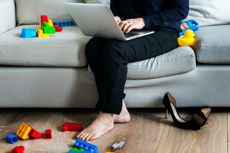 A woman sitting on a couch and typing on a laptop, with children's toys and high heels around her feet