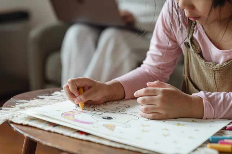 child coloring on a table with parent in background with laptop