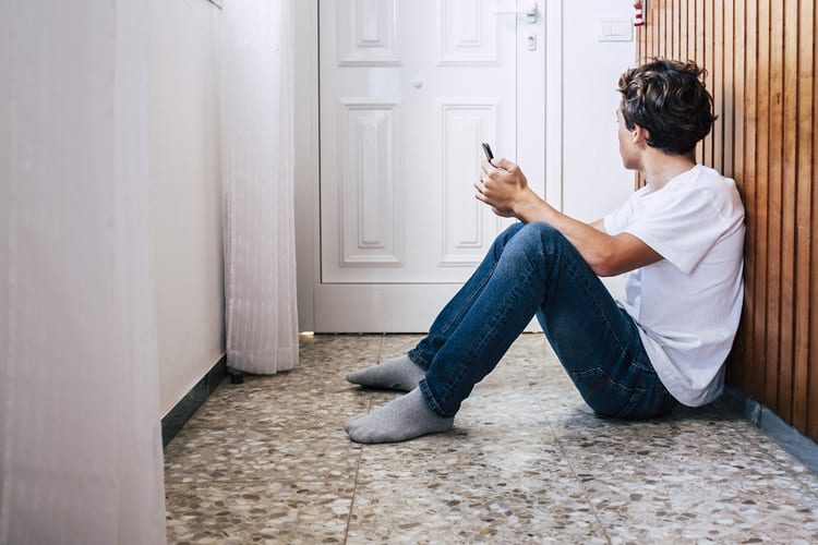 Man sitting on the ground in his home with phone in hand looking at door waiting