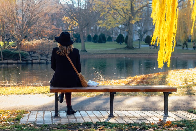 A woman dressed in black coat and hat sits on a park bench overlooking lake in fall season