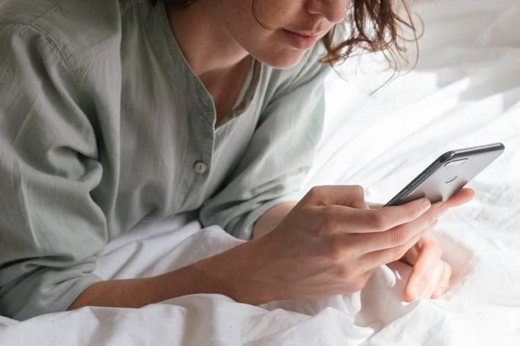 Woman leaning on elbows in bed playing with her iPhone