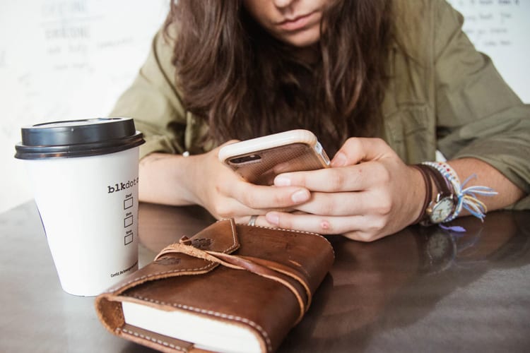 A brunette using her phone, with a coffee cup and journal in the foreground