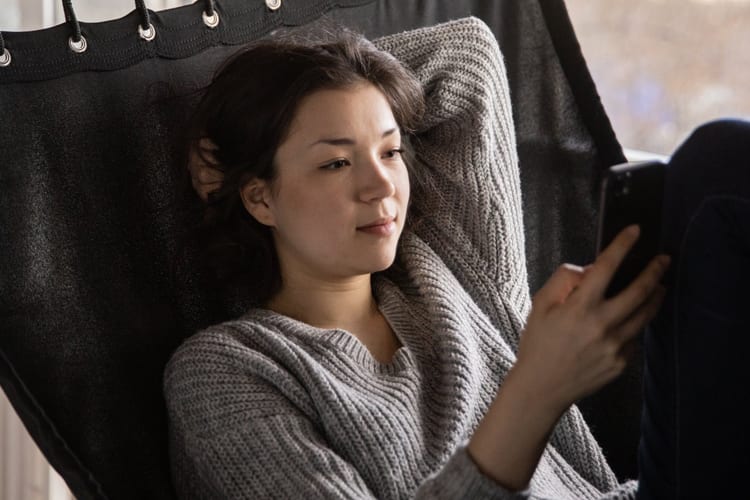 Brunette in a gray cozy sweater relaxing on hammock looking at the phone