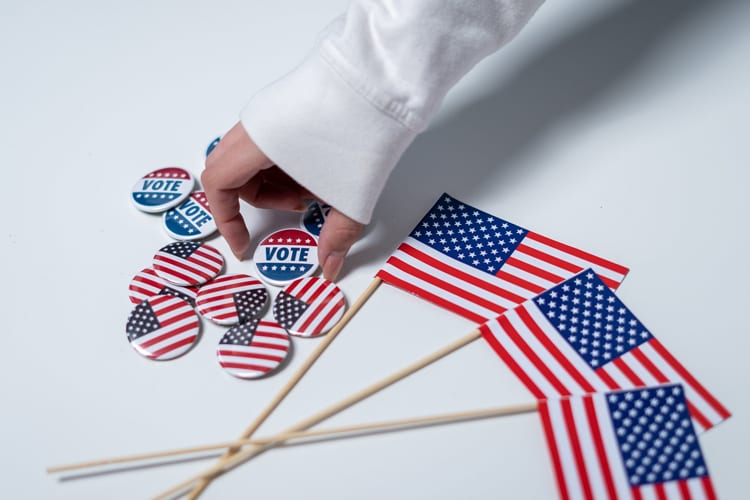 An arm in a white sweatshirt picking up a voting button next to miniature American flags