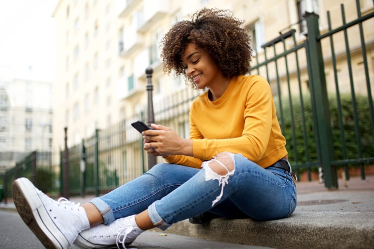 Black woman sitting on curb smiling at smartphone