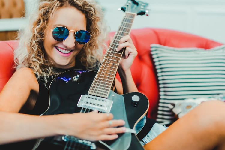 Blonde curly haired woman with blue sunglasses playing guitar on a red couch
