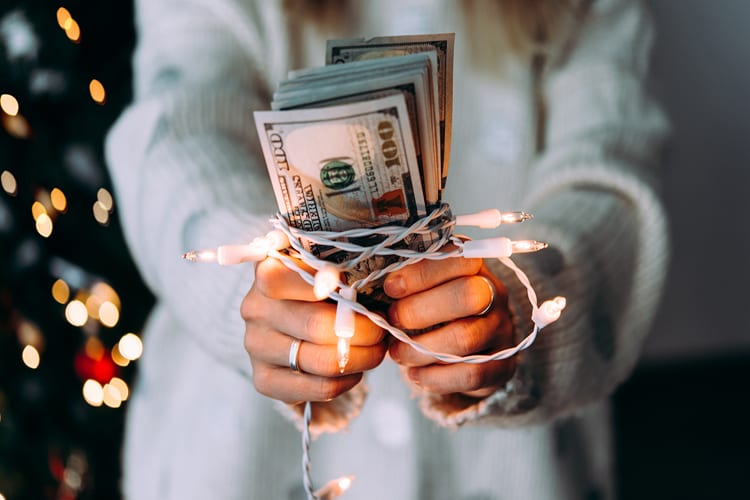 Woman in cozy white sweater holding 100 dollar bills with Christmas lights and tree