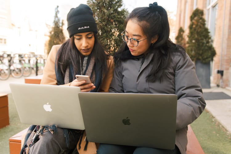 Two women sitting next to each other with Macbooks looking at a phone outside