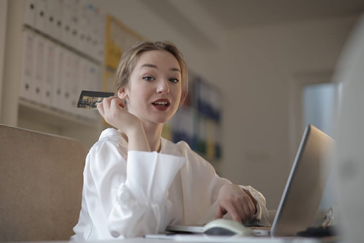 Woman in white blouse holding a credit card looking at camera while on her laptop