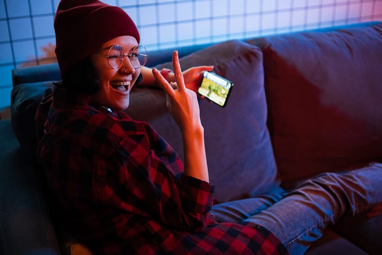 Girl gamer in red flannel and beanie holding peace sign while holding phone with mobile game