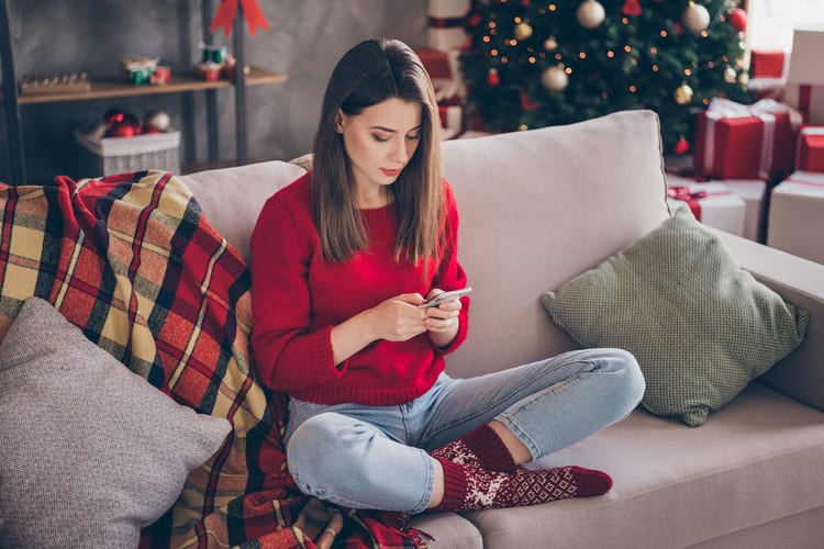 Brunette in red sweater sitting cross-legged on couch looking at phone during holidays