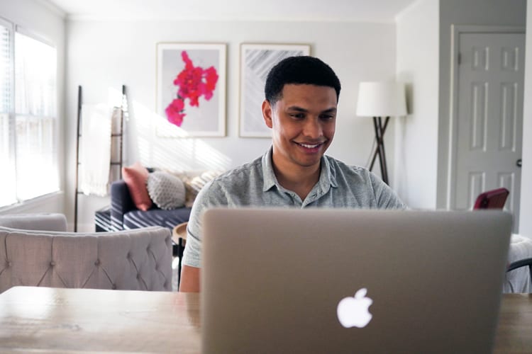 A millennial man sitting in front of a Mac laptop smiling at home
