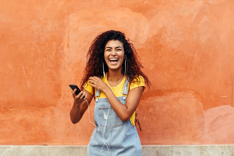 Laughing woman of color listening to music on phone with earbuds on against bright wall
